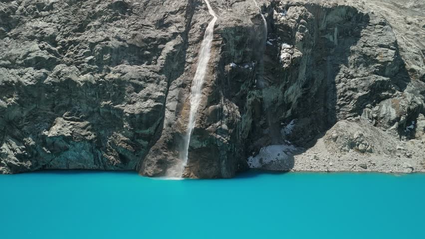 A spectacular aerial parallax shot orbits a majestic waterfall of glacial meltwater as it cascades down a rocky cliff directly into the vibrant turquoise waters of Laguna 69 in Peru.