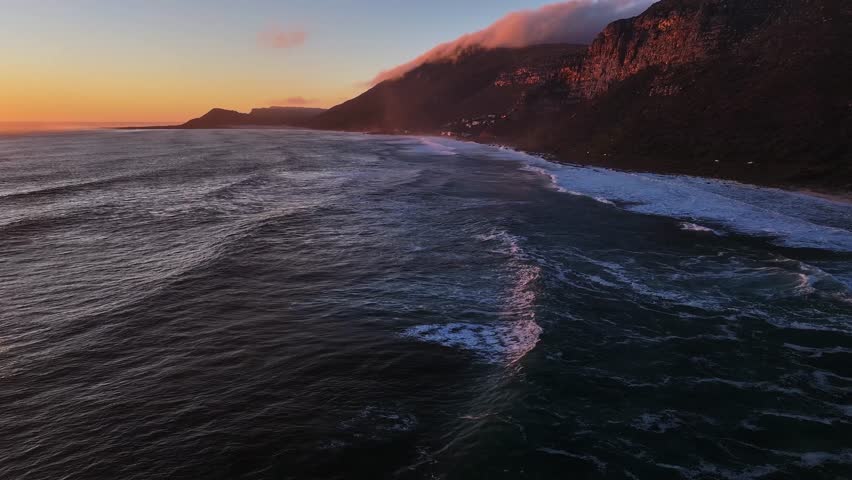 Aerial view of the rugged coastline with dramatic mountains meeting the sea under a vibrant sunset sky, Cape Town, Western Cape, South Africa.