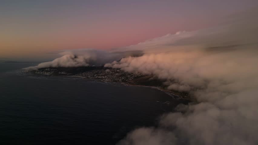 Aerial view of Camps Bay shrouded in white clouds, contrasting with the dark blue ocean and the city lights, Cape Town, Western Cape, South Africa.