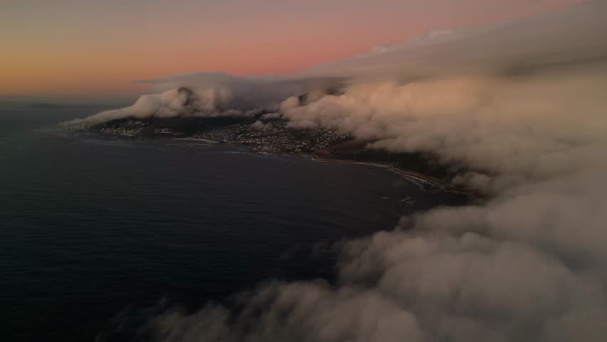Aerial view of a coastal city and coastline shrouded in ethereal clouds, with an early morning sky painted in hues of pink and orange, Cape Town, Western Cape, South Africa.