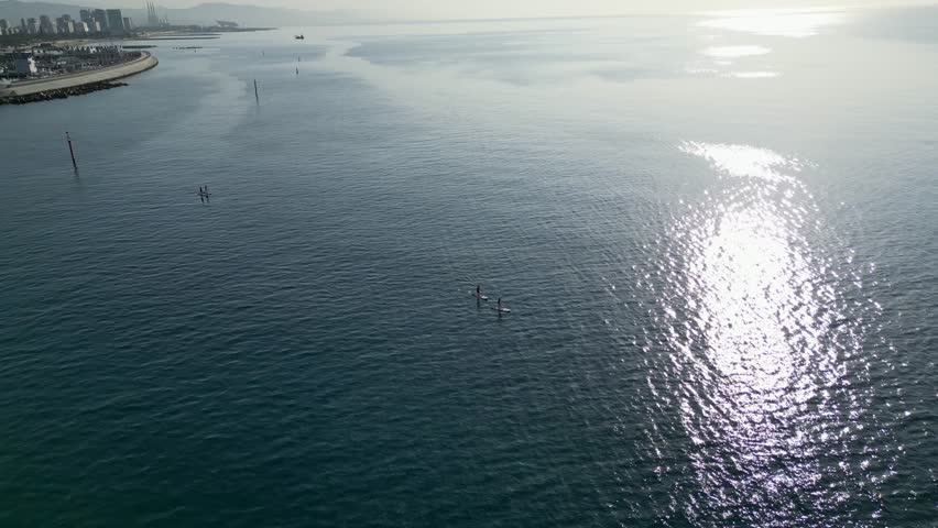 Aerial view of two windsurfers gliding across the turquoise sea near a sun-kissed beach with the city skyline, Barcelona, Catalonia, Spain.