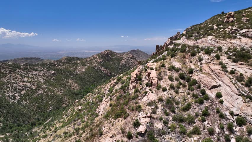 Aerial view of winding road cutting through the rugged terrain, contrasting with the blue sky and the distant city, Santa Catalina Mountains, Arizona, United States.