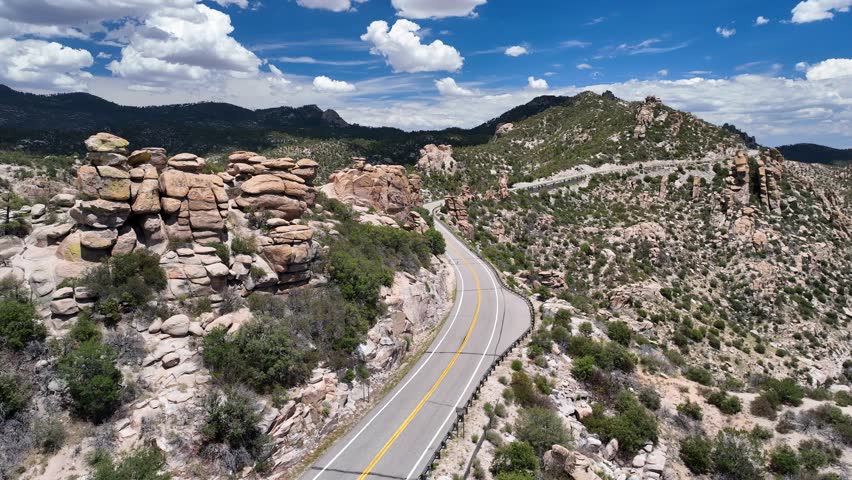 Aerial view of a winding road through the rugged, rocky terrain of mountains under a blue sky with fluffy white clouds, Santa Catalina Mountains, Arizona, United States.