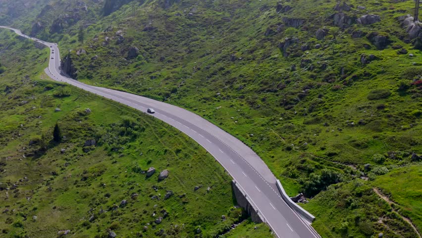 Close aerial shot of two cars cruising along a smooth mountain road flanked by alpine greenery near Hospental