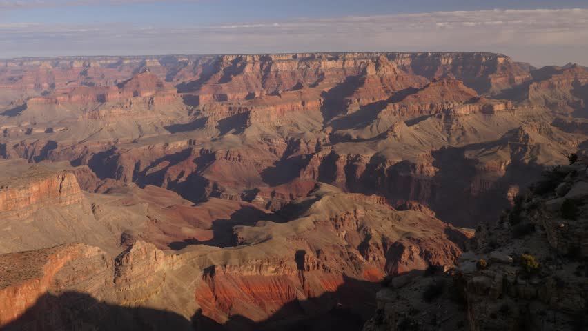 Experience a stunning aerial view of the majestic Grand Canyon, showcasing dramatic rock formations bathed in warm sunlight. Timelapse captures the changing hues of this awe-inspiring natural wonder.