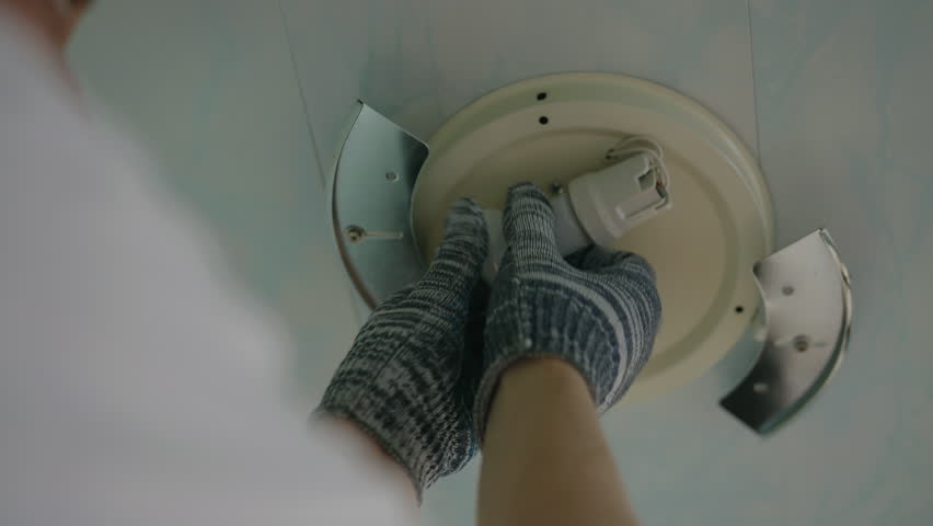 Close-up hands of unrecognizable electrician wearing gloves replacing light bulb in ceiling light fixture at home. Concept of electricity maintenance, proper illumination and electrical safety.