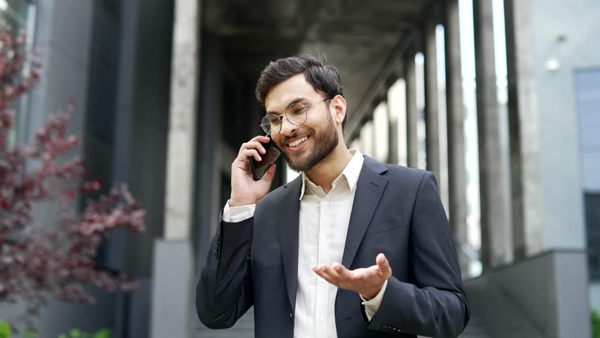 Smiling businessman in formal suit talking on mobile phone standing on street near office building. Happy professional male entrepreneur communicates with business partner or client on smartphone