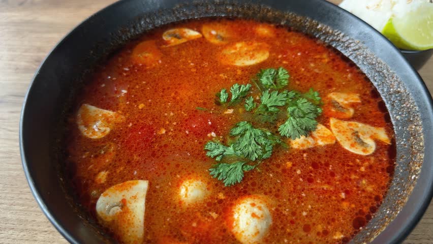 Hot spicy red Thai soup Tom yum with shrimps and mushrooms, served with cilantro and fresh lime in black bowl on a wooden table, top view while moving up
