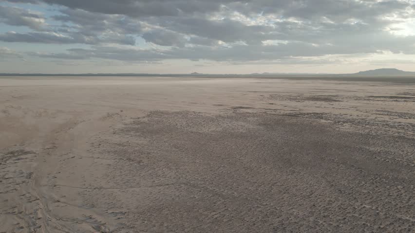 Aerial drone footage of a dried-up lake with sandy ground scattered with small bushes, under a clear blue sky and hills in the distance