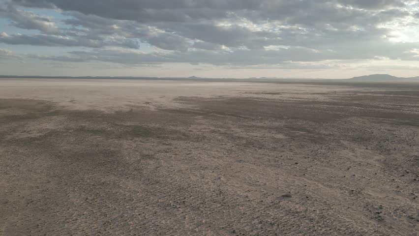 Aerial drone footage of a dried-up lake with sandy ground scattered with small bushes, under a clear blue sky and hills in the distance