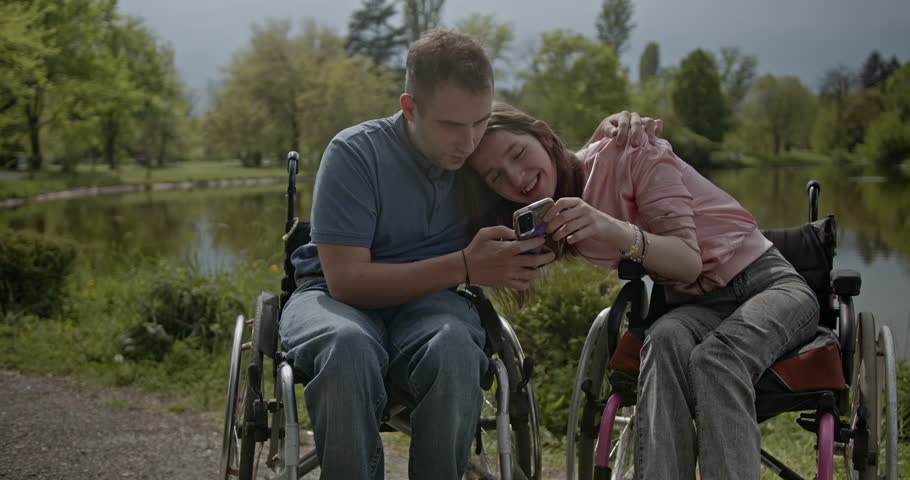 A happy and inclusive couple in wheelchairs shares a joyful moment, looking at a mobile phone together in a beautiful park by the lake. They are smiling, showing connection and modern leisure.