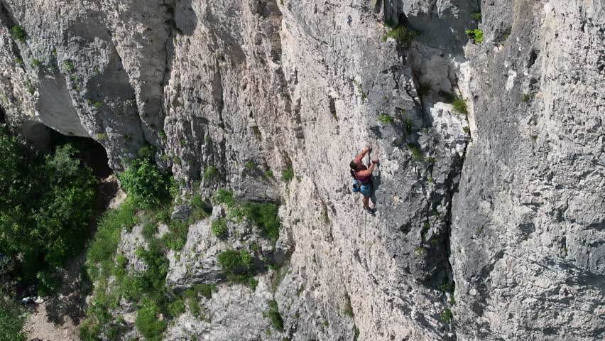 AERIAL: Female rock climber lead climbing a steep rock face on a sunny day. Scenic vertical wall is surrounded by lush greenery. Challenging adrenaline sports activity in beautiful natural environment