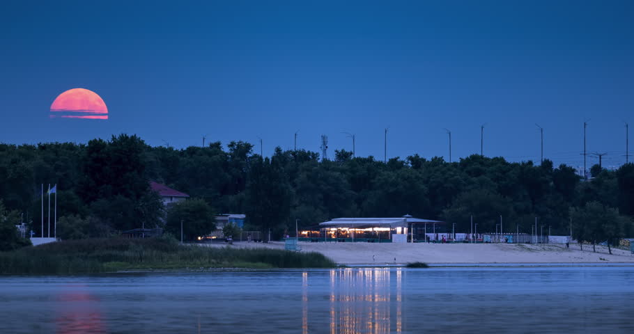 Full moon rising in August over the Dnipro River in Kyiv, Ukraine, casting warm reflections on the water, with a beach and illuminated building under the deep blue evening sky. Time lapse
