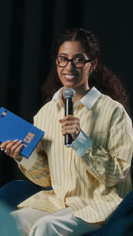 Vertical of young biracial female author holding her latest book speaking into microphone on stage in front of audience