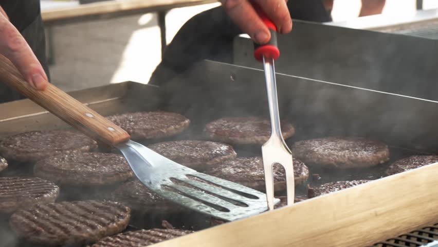 detail of hands carefully checking the cooking of many hamburgers on a large griddle surrounded by smoke