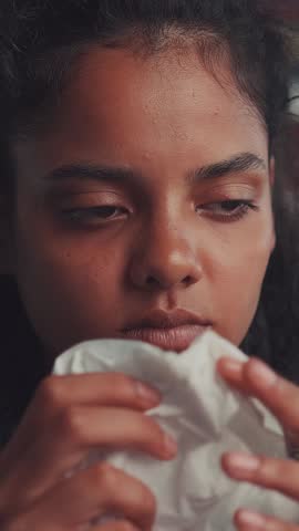 A young Indian woman, feeling under the weather with a cold or flu, blows her nose into a wet tissue and rubs her temples, struggling with discomfort and mild pain from her illness.