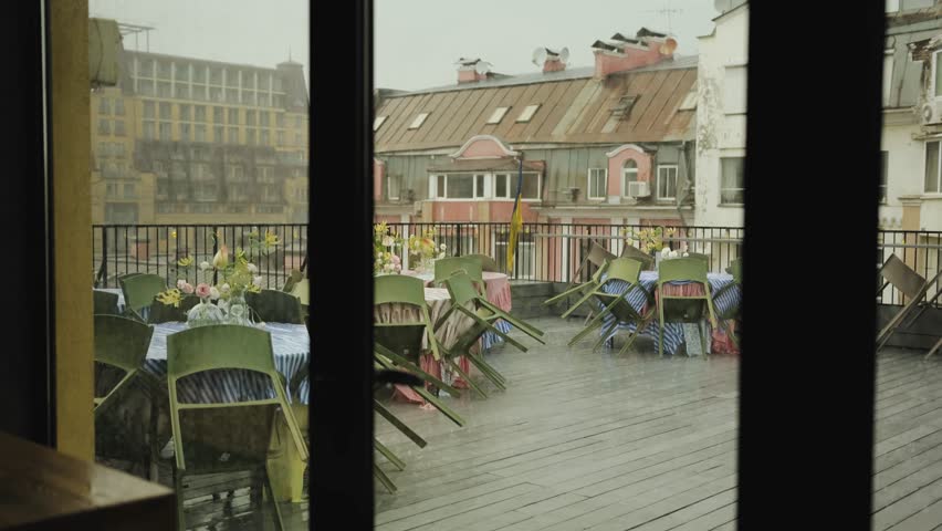 View through sliding doors onto a terrace of a cafe or restaurant where it is raining. Empty wet tables, rain falling on the wooden floor. Slow motion. The restaurant is closed due to bad weather.