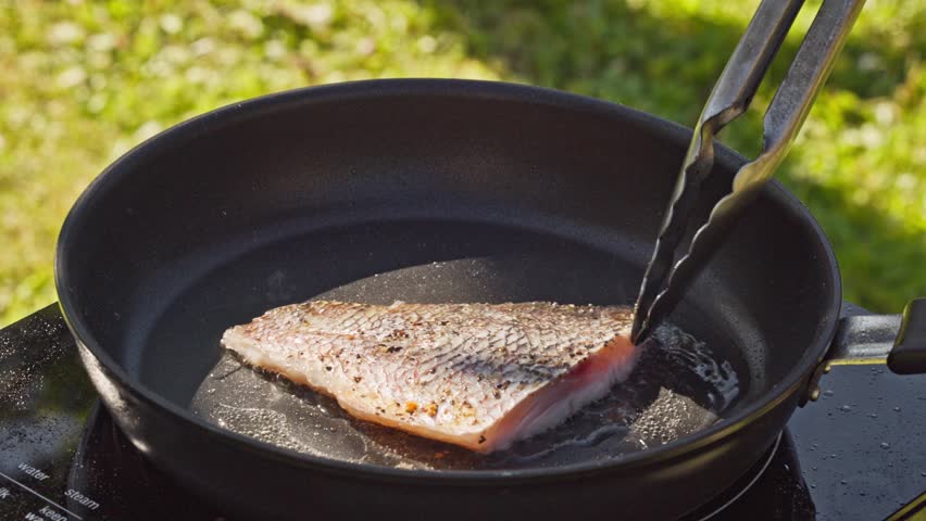 outdoor cooking. cook preparing fresh fish fillet on hot pan with oil for making delicious meal in garden, food closeup, outdoors natural light