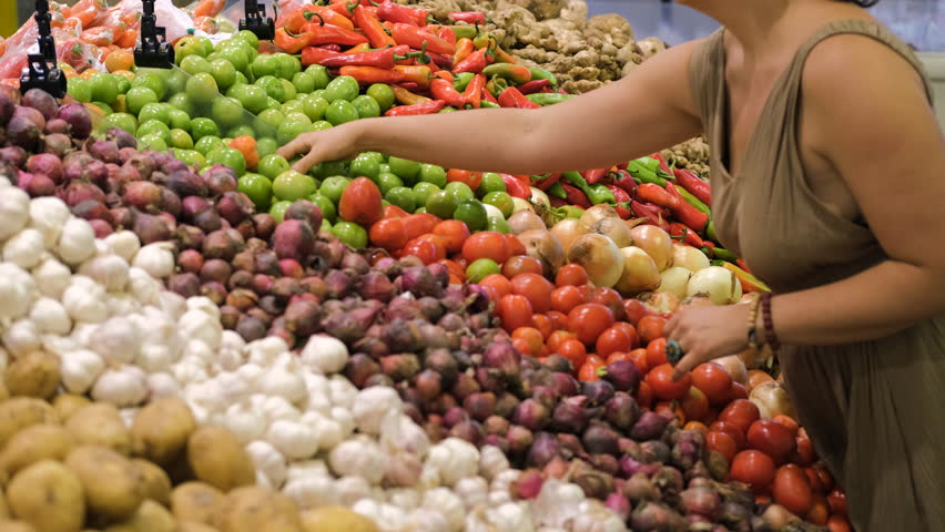 Woman selecting green tomatoes among a vibrant display of fresh vegan produce in a grocery store. She chooses organic vegetables for her meals, emphasizing healthy lifestyle, slow motion