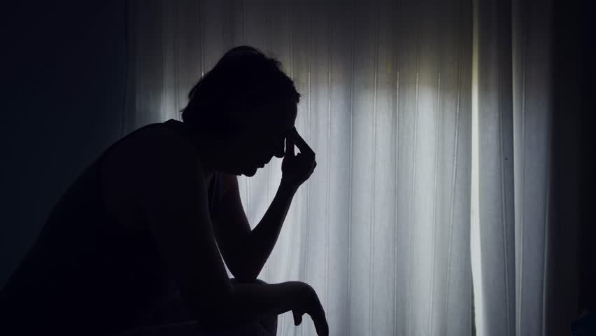 Silhouette of a woman sitting by a window, holding her head in distress, expressing anxiety, sadness, or emotional struggle. 4K with selective focus.