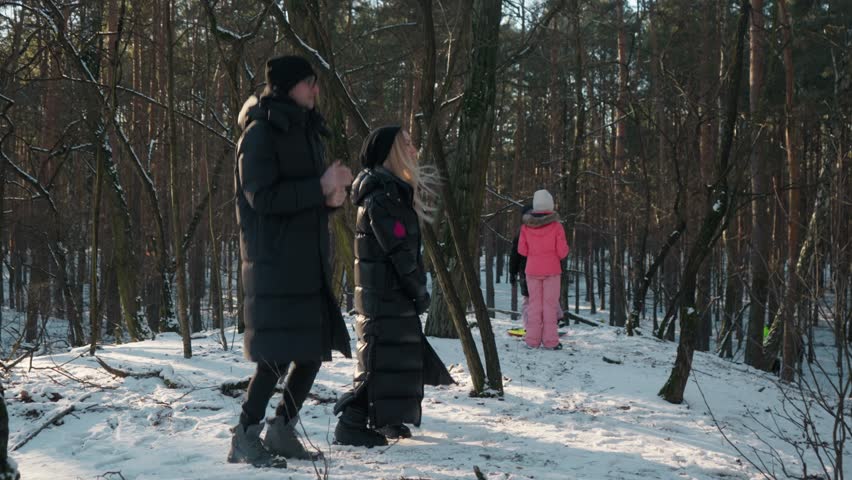 A young couple feels cold during a winter walk in a snowy forest. A man and a woman are jumping and rubbing their hands, trying to get warm while watching their children play in the frosty weather.