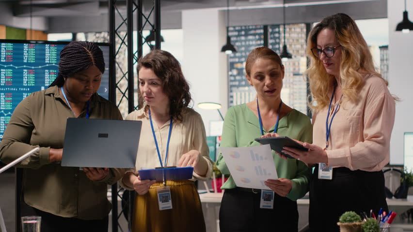 Ambitious and goal oriented diverse businesswomen reviewing corporate reports and graphs, working in synergy to achieve professional growth in a modern work environment. Camera B.