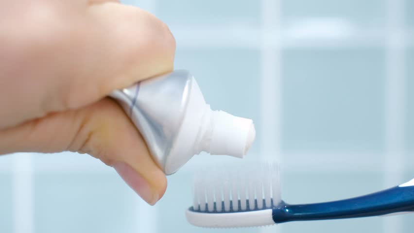 Female hand squeezing toothpaste onto toothbrush against blue tile background.
