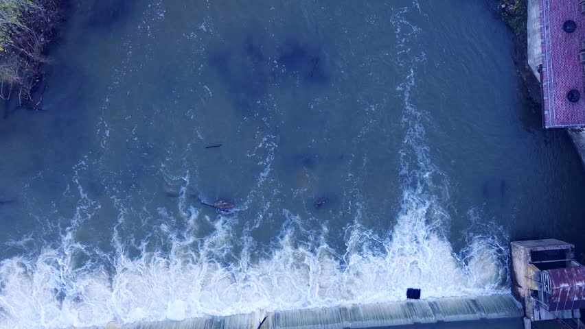 Drone panning over a dam then following the river flow to a lit footbridge surrounded by vibrant yellow and orange trees and lush green grass in scenic Missouri fall HD footage at golden hour