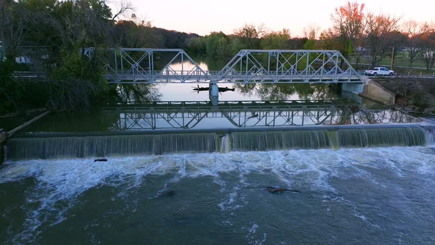 Drone flying above a river in Missouri toward a dam and bridge before rising over and passing the bridge in smooth cinematic HD footage during golden hour with peaceful scenic views