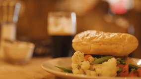 A hand uses knife and fork to cut a golden pie served with steamed vegetables on a plate, under warm ambient lighting in a cozy pub environment - Powered by Shutterstock - Get 15% off with code: PIKWIZARD15