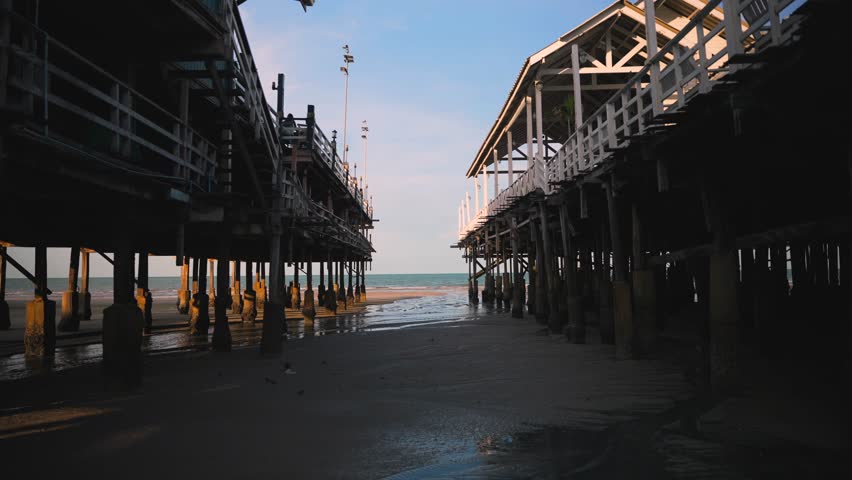 Wide view between two wooden piers on stilts, revealing wet sand and the distant blue ocean at sunset during low tide.