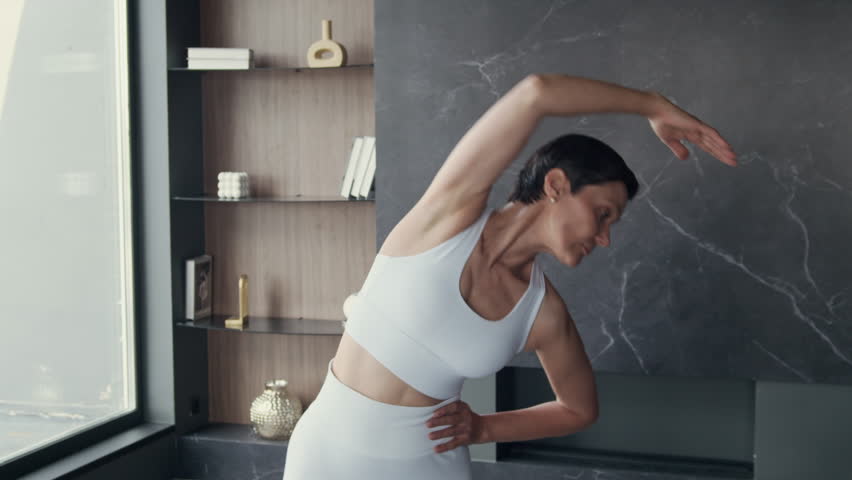 Medium copy space shot of middle-aged Caucasian woman wearing white activewear stretching body parts in minimalist furnished apartment