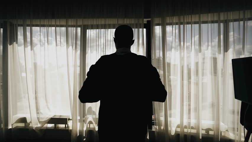The man puts on his jacket as he approaches the large window. his silhouette looks expressive in the frame. Shooting a man from behind