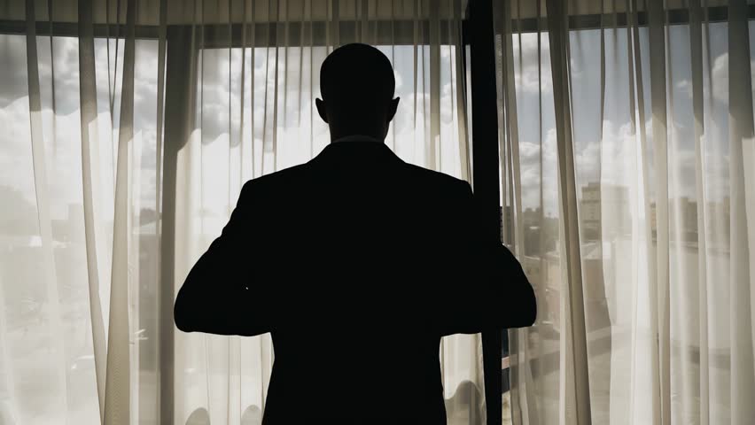The man puts on his jacket as he approaches the large window. his silhouette looks expressive in the frame. Shooting a man from behind