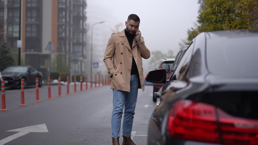 A very fashionable man dressed in a stylish trench coat holds a phone while standing near parked cars on a foggy street