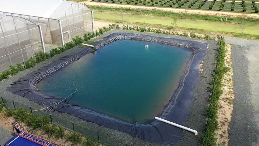 A drone shot of covered water reservoir beside greenhouse facility, used for farm irrigation and water storage