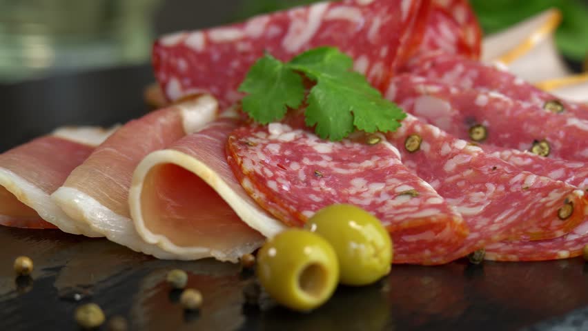 Close up of fresh slices cold cuts sausage ham bacon jamon rotate in circle on stone cutting board with olives and parsley. Meat. Pork beef. Food