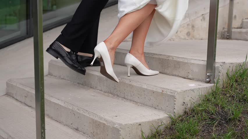 A Close-up slow motion of a bride in white heels and groom in black shoes walking up concrete stairs in elegant wedding outfits