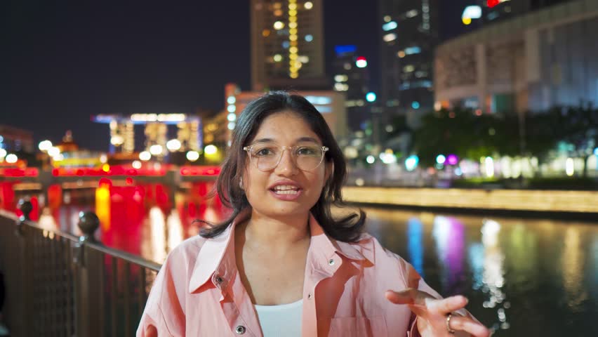 4K Portrait of confident Indian woman vlogging at Clarke Quay during night at Singapore city. Stylish female tourist filming a video and introducing tourist attractions. Holidays and travel concept.