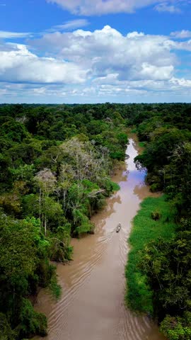 A vertical drone shot of a narrow Amazon tributary winding through rainforest in Peru, with a canoe leaving soft ripples under a dramatic cloudscape