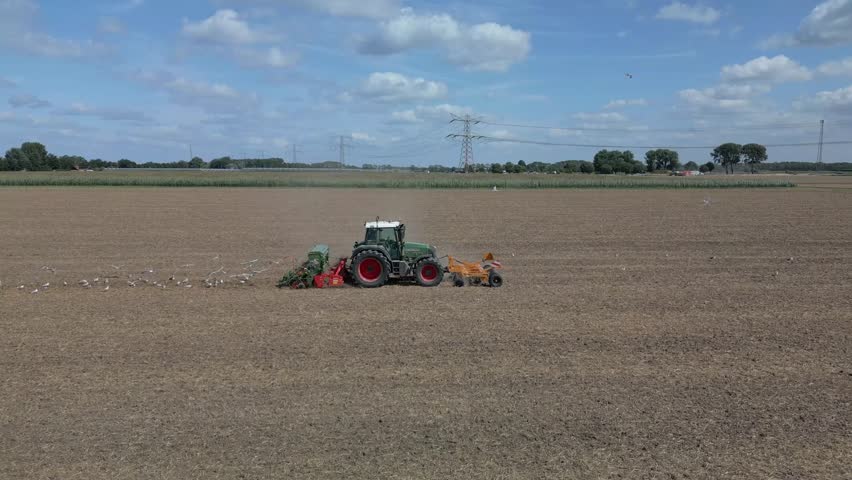 A tractor tilling dry farmland with seagulls trailing, under a bright sky with scattered clouds