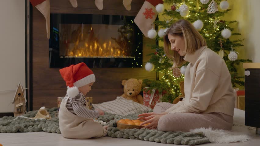 A little boy in a festive hat and his mother share a joyful moment playing with oranges beside a beautifully decorated Christmas tree.