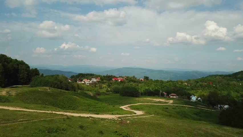 A drone view of dirt road winds through grassy hill towards a small village. The sky is full of white clouds, and a mountain range is in the distance.