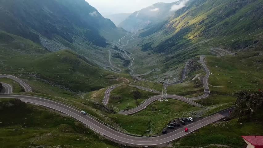 A drone view of Transfagarasan Road winds through a lush, green valley, surrounded by the Carpathian Mountains with misty horizon in Romania