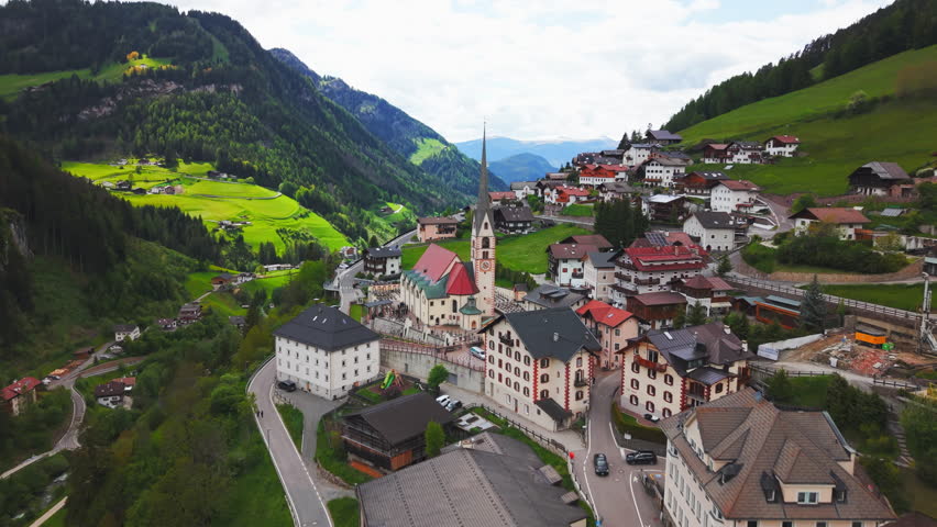Drone footage of a slow rising aerial shot transitioning to a top-down view of a church with red roof and surrounding alpine village in the Dolomites, Italy.