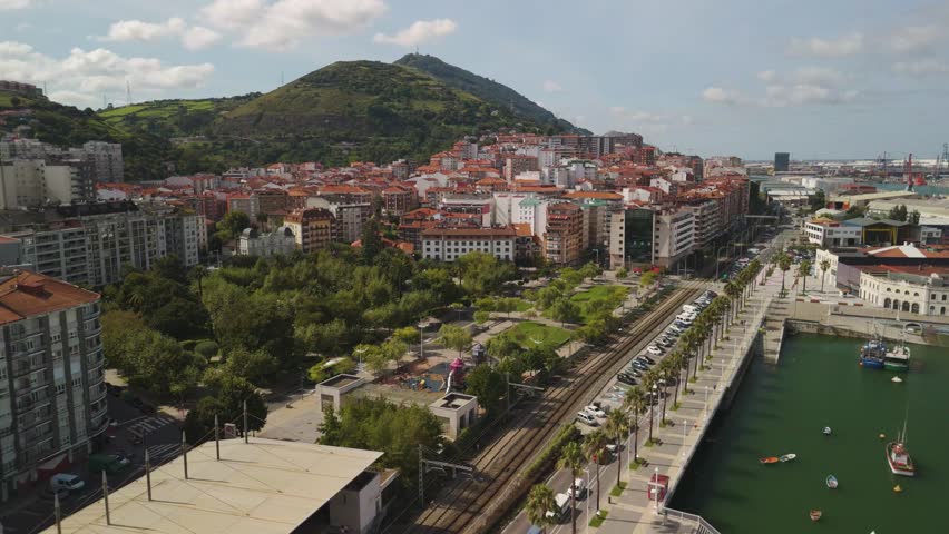 A drone view of Bilbao city nestled at the base of a green hill. A road with palm trees lines a body of water with small boats in Spain