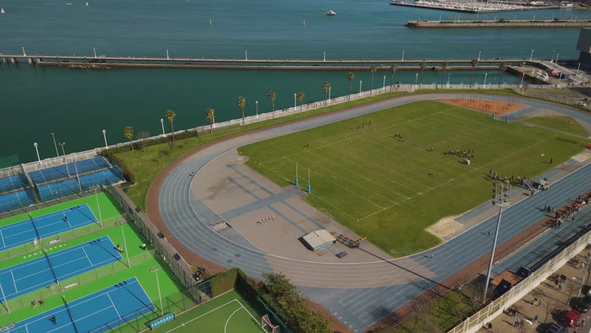 An aerial view of a coastal sports complex with a track, American football field, and tennis courts surrounded by water and some buildings in Spain