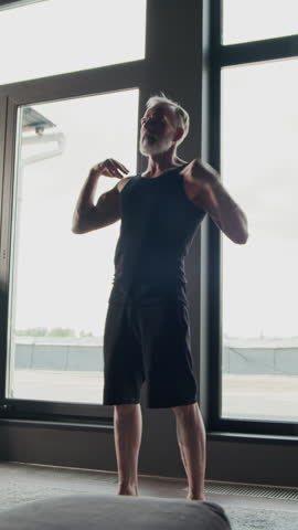 Vertical shot of adult Caucasian gray-haired man in black activewear practicing shoulder circles exercise during home workout