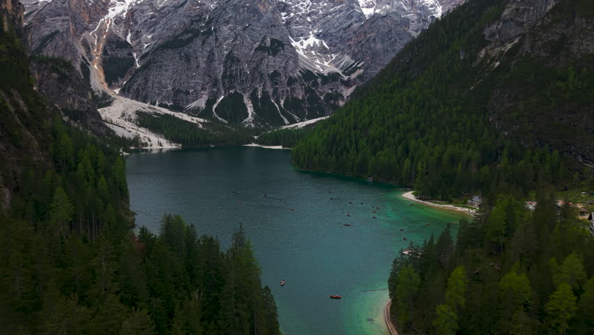 Aerial view approaching the shore of Lago di Braies with turquoise water, boats, and pine forest in the Dolomites, Italy.