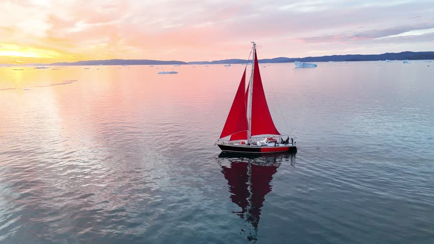 Little red sailboat cruising among floating icebergs in Disko Bay glacier during midnight sun season of polar summer. Ilulissat, Greenland. Global warming and melting glaciers.
